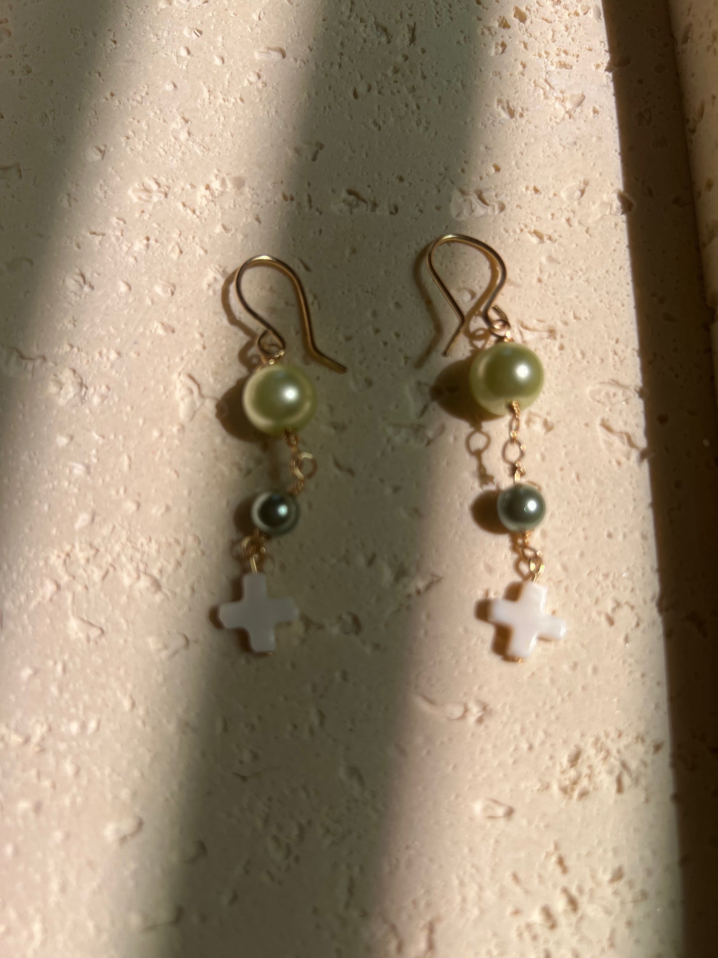 Close-up of earrings with light green and dark teal pearl beads and white cross pendant on gold wire