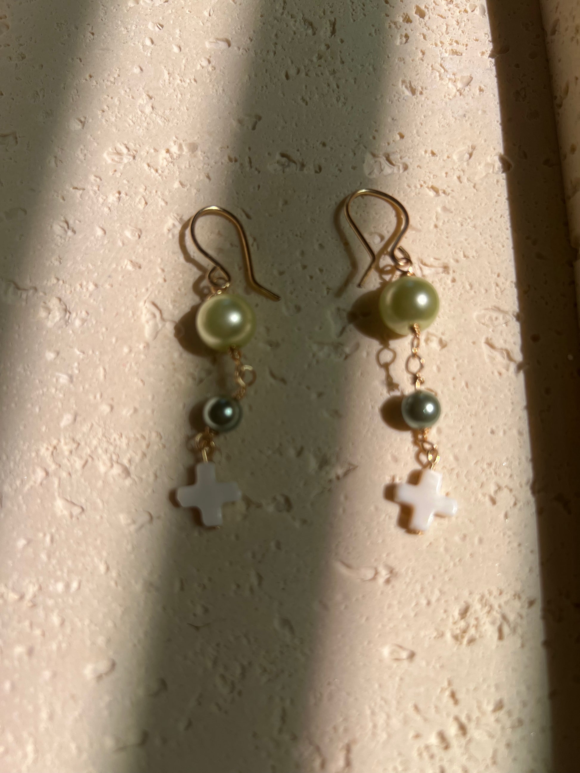 Close-up of earrings with light green and dark teal pearl beads and white cross pendant on gold wire