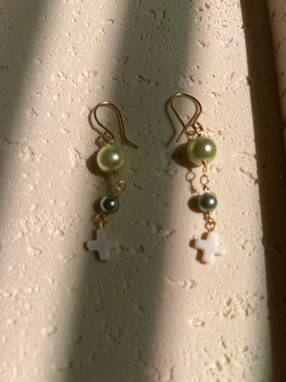 Close-up of earrings with light green and dark teal pearl beads and white cross pendant on gold wire