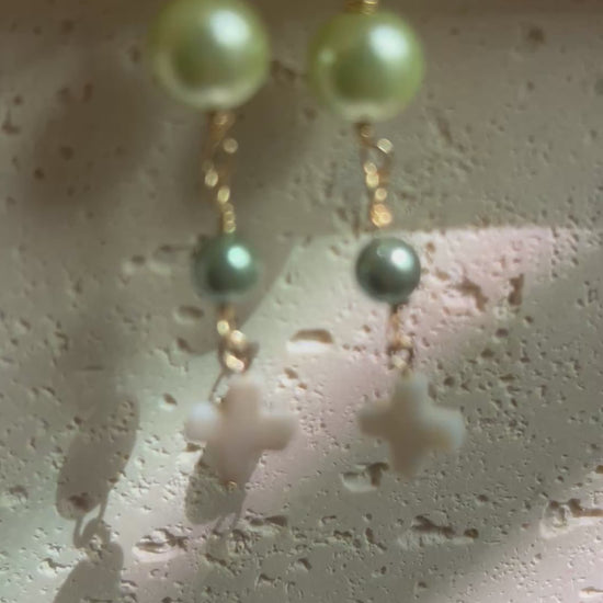 Earrings held in hand showing light green and dark teal pearl beads with white cross drop on gold hooks