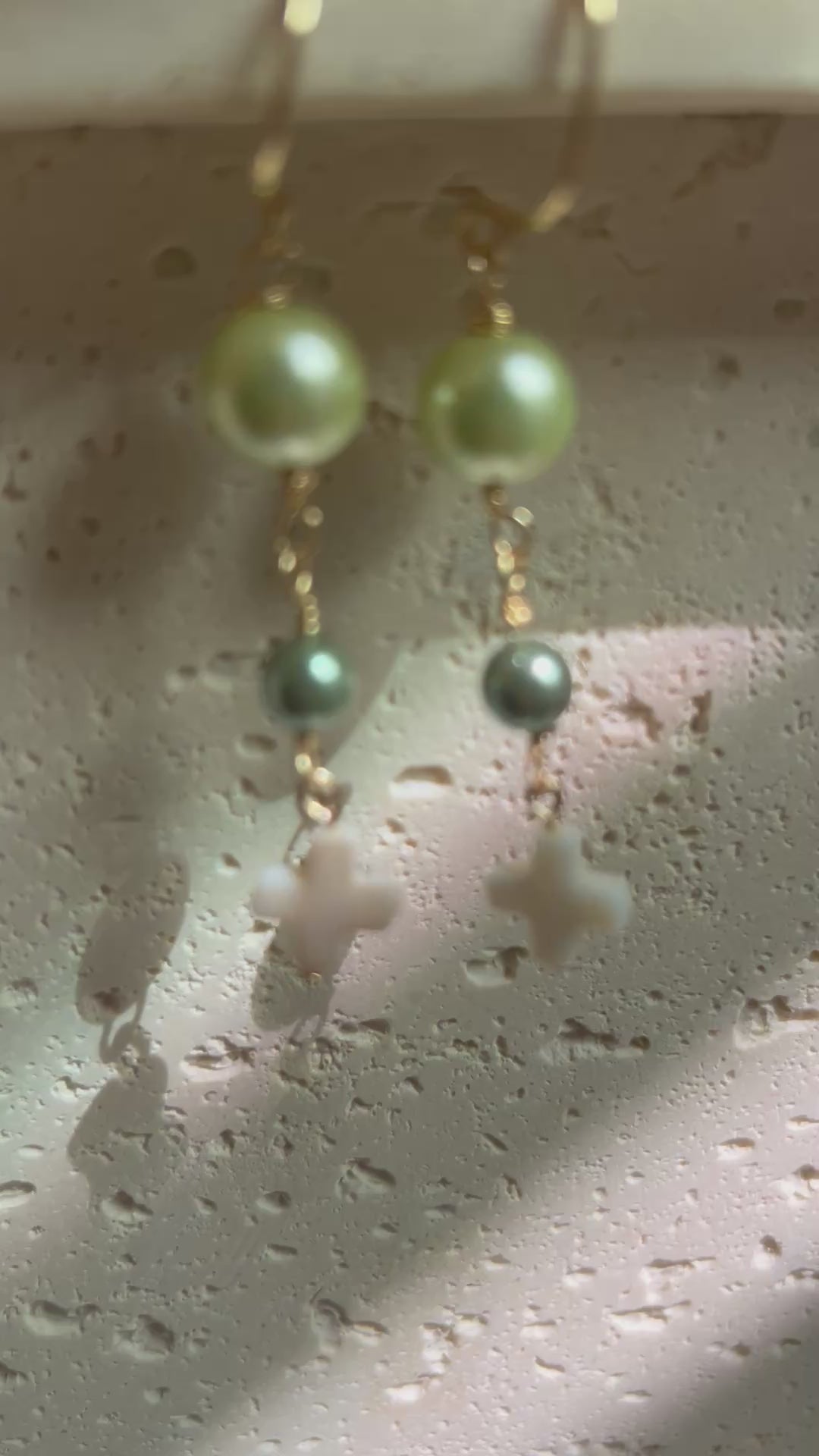 Earrings held in hand showing light green and dark teal pearl beads with white cross drop on gold hooks